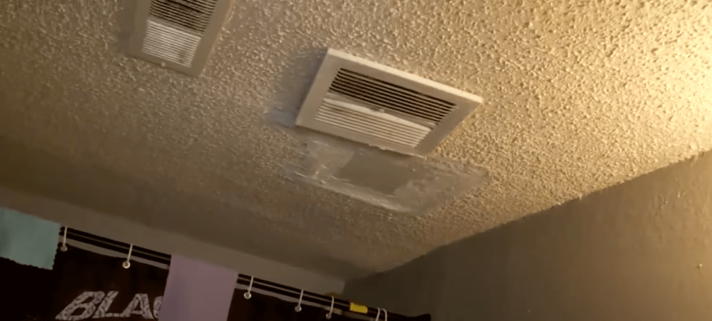 a bathroom ceiling with two ventilation grilles installed. The ceiling has a textured (popcorn) finish, and there is visible water damage or a poorly patched area around one of the vents. The patch looks like it was done to cover a previous leak or mold-affected section, as it's discolored and uneven compared to the surrounding ceiling.